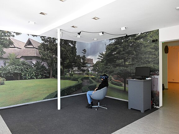 Ein Galerieraum mit Teppichboden, an den Wänden Fototapete mit einer aufgedruckten Parklandschaft. In der Mitte des Raums befindet sich ein Stuhl, in dem man sich mit einer VR-Brille in eine andere Welt begeben kann.