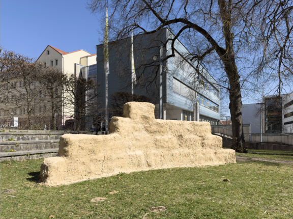 Eine Skulptur der Künstlerin Syowia Kyambi. Die Skulptur besteht aus Strohballen, die mit einer Mischung aus Kuhdung und Erde verputzt wurde. Die Skulptur steht auf der Wiese im Hinterhof der Galerie. Im Hintergrund sind ist ein Gebäude der Hochschule Furtwangen und ein blauer Himmel ohne Wolken sichtbar. Die Umgebung wirkt offen und urban.