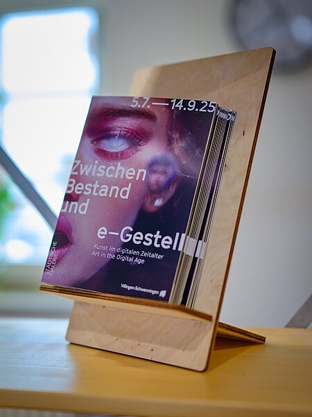 Ein Aufsteller mit einem Flyer, der für eine Kunstausstellung mit dem Titel "Zwischen Bestand und e-Gestell" wirbt. Die Ausstellung findet vom 5. Juli bis 14. September 2025 statt und thematisiert Kunst im digitalen Zeitalter.