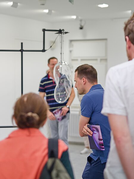Die Szene zeigt eine Kunstinstallation in einem weißen Raum. Mehrere Personen beobachten eine transparente, organisch geformte Skulptur, die von einem Gestell hängt. Einige Teilnehmer halten Bilder in der Hand, während sie sich auf das Kunstwerk konzentrieren.