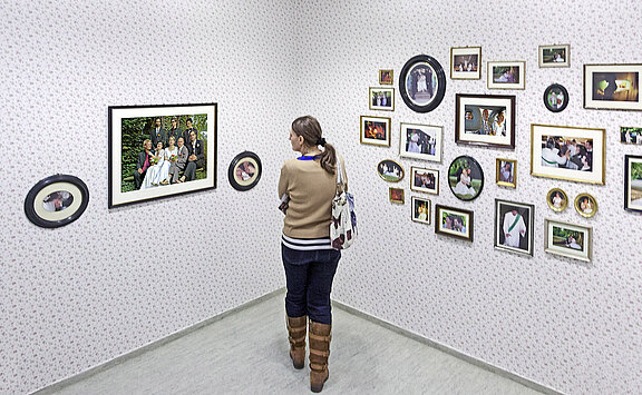 Eine Person steht in einem Raum, schaut sich ein großes Familienfoto an, das eine Gruppe von Menschen in festlicher Kleidung zeigt. Um sie herum hängen zahlreiche weitere Bilder in verschiedenen Rahmen an der Wand. Der Raum hat eine blumige Tapete.