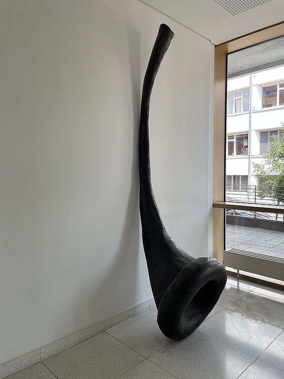 Große schwarze, gebogene Skulptur in Horn- oder Trompetenform: langes, schlankes Rohr mit weitem, geschwungenem Trichter am Fuß, angelehnt an eine weiße Wand neben einem Fenster.