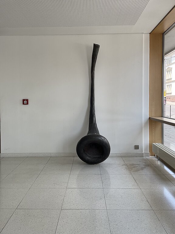 Andrea Zaumseil Langhalsige, schwarze Skulptur mit trichterartiger, schalenförmiger Basis, hochkant an einer weißen Wand platziert.