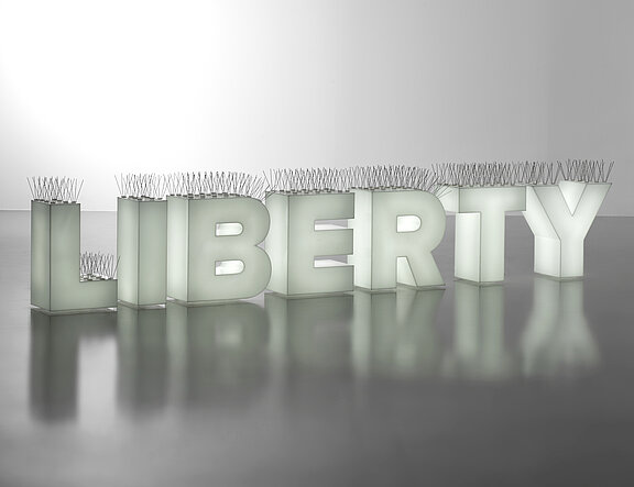 Leuchtende Großbuchstaben bilden das Wort „LIBERTY“, auf deren Oberseiten schmale Metallstacheln zur Vogelabwehr angebracht sind; die Schrift spiegelt sich auf dem glatten Boden.