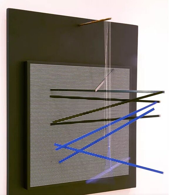 Abstrakte Wandskulptur: schwarzes Rechteck mit feinem, horizontalem Weißlinienfeld, darüber gekreuzte diagonale Stäbe in Schwarz und Blau, die eine geschichtete optische Komposition bilden.