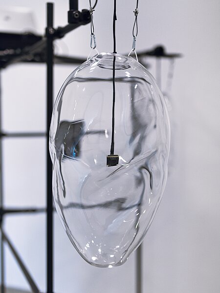 Eine transparente, gläserne Skulptur in Form eines Gesichts hängt an Drähten. Der Gesichtsausdruck ist abstrakt und fließend, mit sanften Kurven und Linien, die das Licht reflektieren. Im Hintergrund sind unscharfe Strukturen zu erkennen, die die künstlerische Umgebung unterstützen.