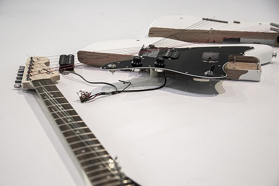 Das Bild zeigt mehrere elektrische Gitarren, die teilweise zerlegt sind. Eine Gitarre hat einen schwarzen Korpus mit zwei Drehknöpfen und sichtbaren Saiten. Die anderen Gitarren haben helle Holzteile und sind auf einer neutralen Oberfläche angeordnet.