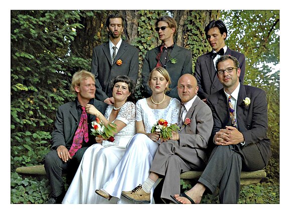 Oberkofler, Gabriela, Hochzeit Eine Gruppe von acht Personen posiert in einem Garten. Zwei Frauen tragen weiße Kleider, die anderen tragen Anzüge. Alle halten Blumensträuße. Die Szenerie ist freundlich und festlich, umgeben von Pflanzen. Die Atmosphäre deutet auf einen besonderen Anlass hin, möglicherweise eine Hochzeit.