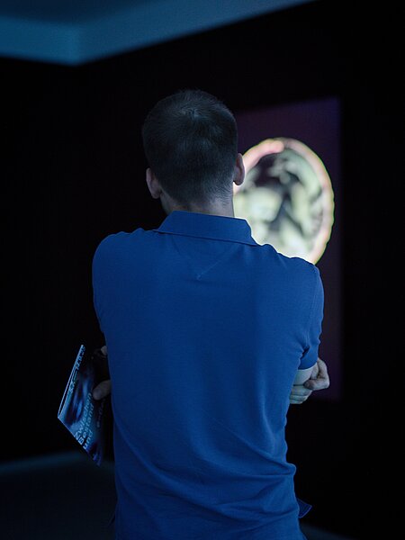 Eine Person mit einem blauen Polo-Shirt steht mit verschränkten Armen vor einem Bildschirm in einem dunklen Raum. Der Bildschirm zeigt eine grafische Darstellung oder ein Kunstwerk.