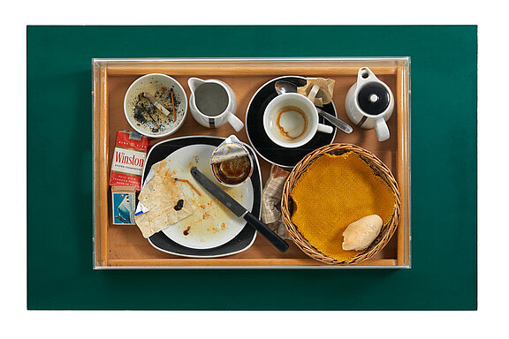 Ein Tablett mit benutztem Geschirr zeigt Reste von Essen und Getränken. Darunter sind leere Tassen, eine Plastiktüte, eine Zigarettenschachtel und ein Brotkorb. Die Oberfläche ist unordentlich und hebt den Eindruck eines gerade beendeten Essens hervor.